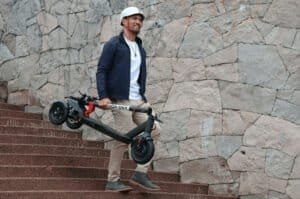 Un homme transporte sa trottinette à trois roues, qui selon Pièces 2 Trott fait partie des différents types de trottinettes électriques à connaître.