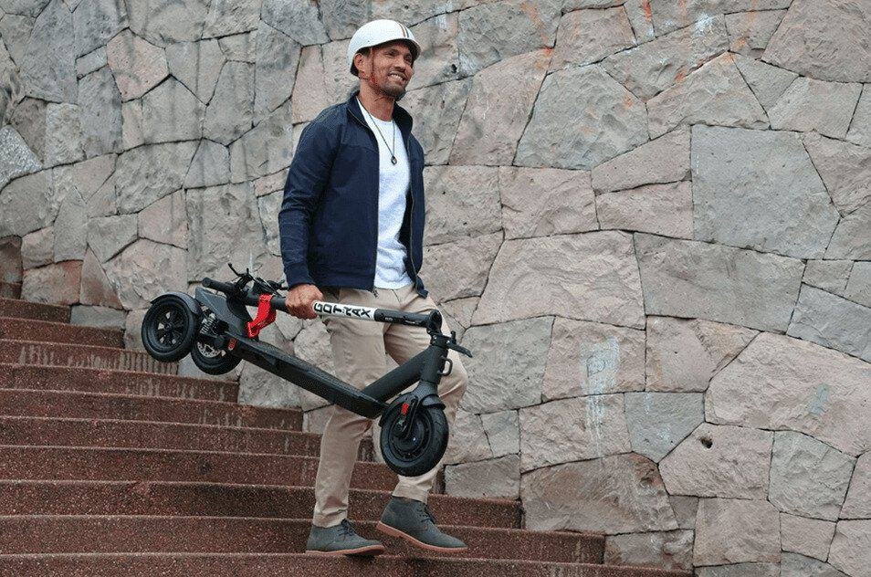 Un homme transporte sa trottinette à trois roues, qui selon Pièces 2 Trott fait partie des différents types de trottinettes électriques à connaître.